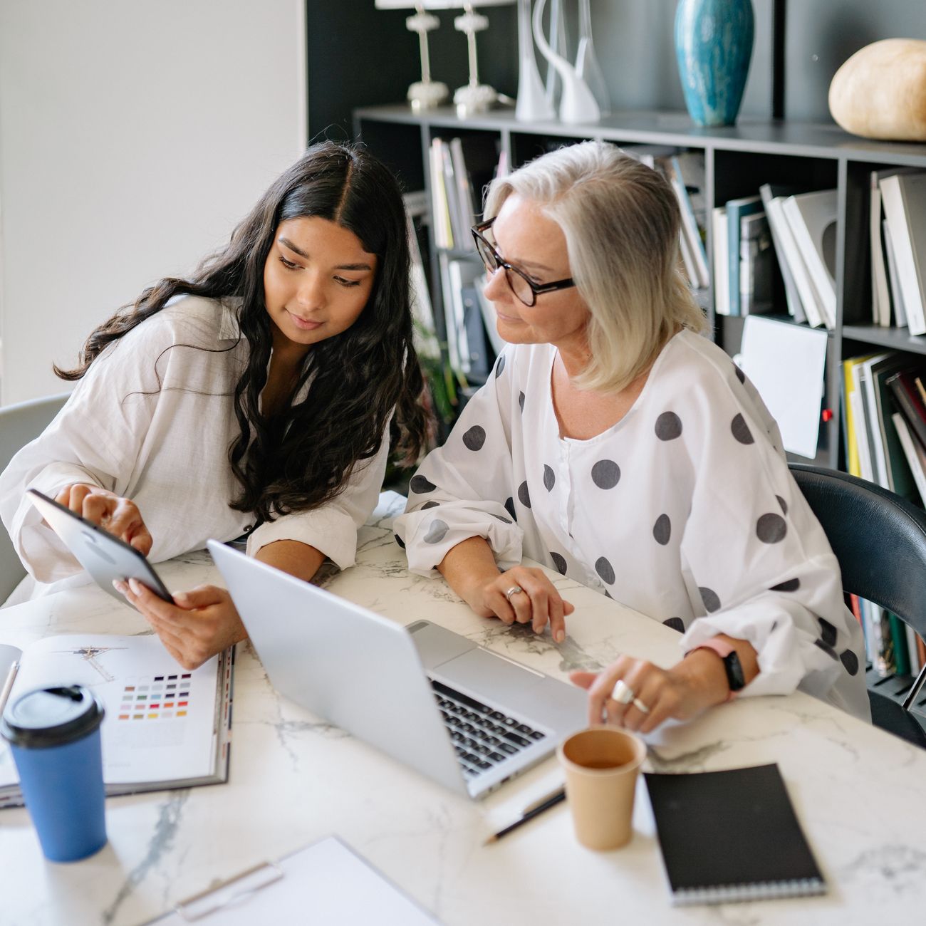 Two women working together at a desk with a laptop and a tablet in an office