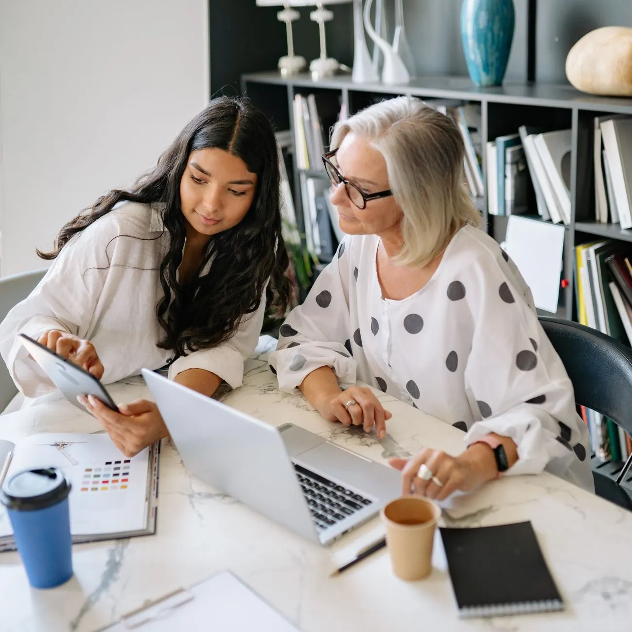 Two women working together at a desk with a laptop and a tablet in an office