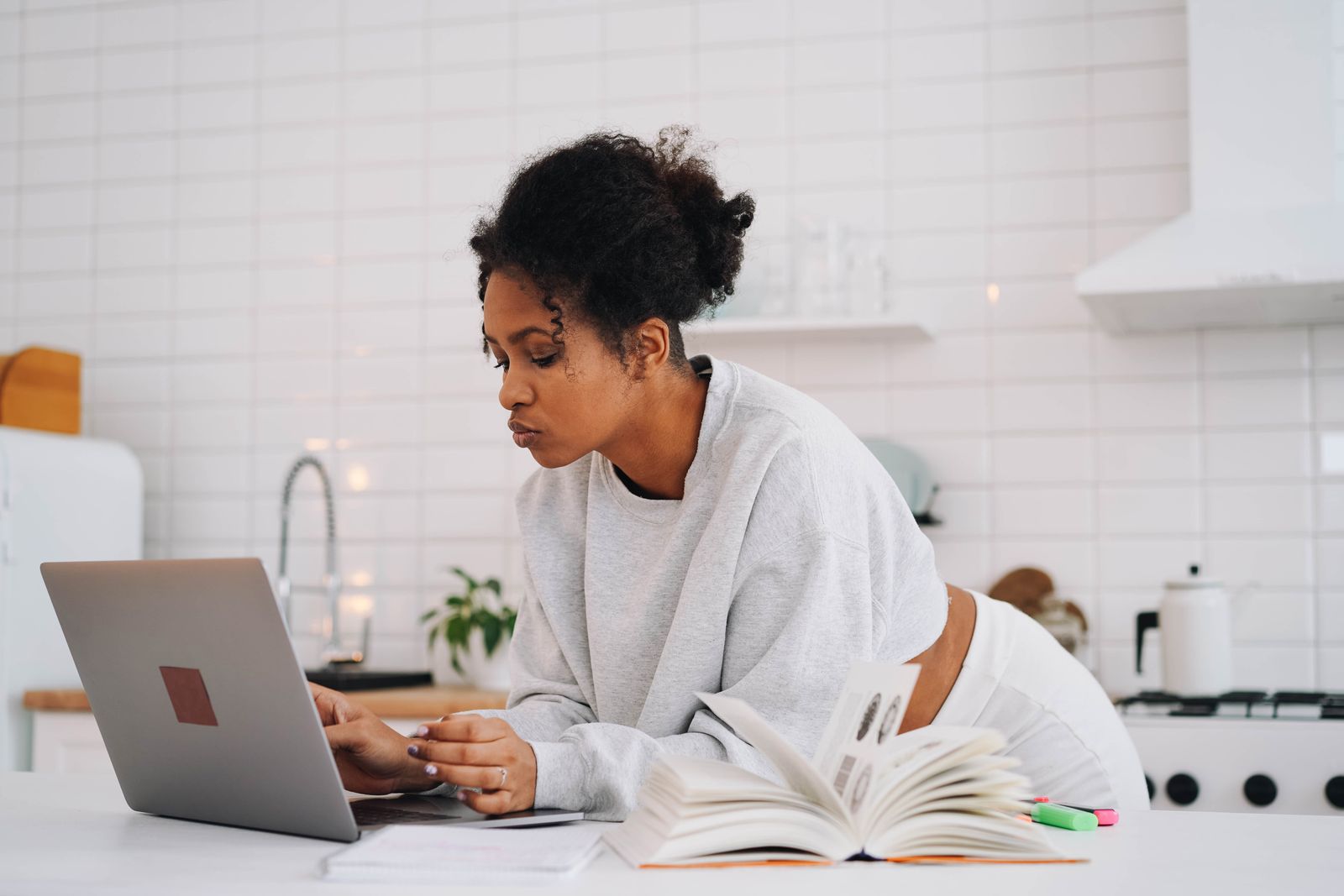 A woman in a white sweater studying on a laptop at her kitchen table with textbooks open beside her