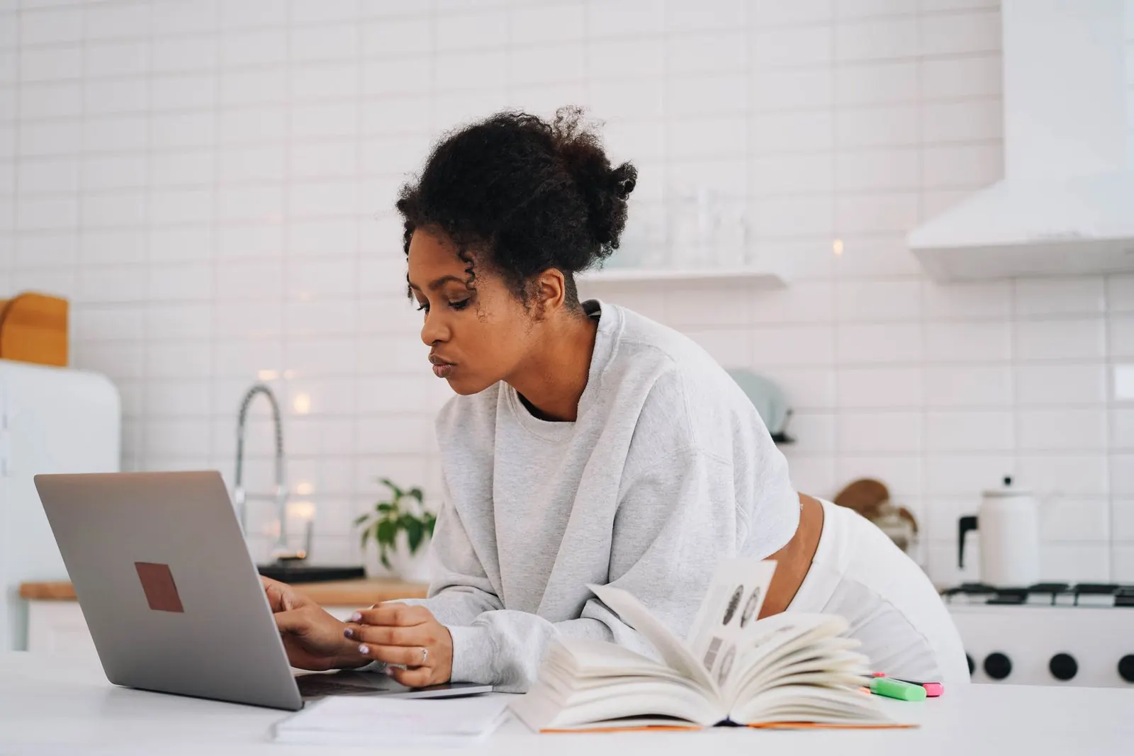 A woman in a white sweater studying on a laptop at her kitchen table with textbooks open beside her